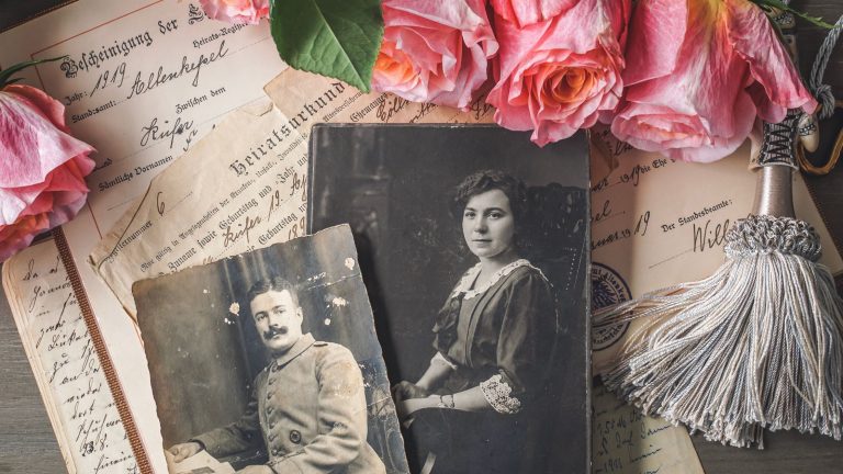 Flatlay of vintage family photos and old documents with pink roses and a tassel, symbolising ancestor connection and ritual honouring of roots.